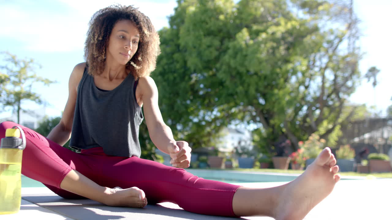 feliz mujer biracial practicando yoga sentada junto a la piscina en el jardín soleado, espacio de copia, cámara lenta