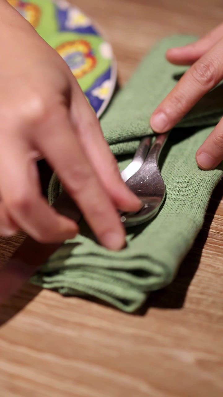 Mãos colocam cuidadosamente faca, colher e garfo sobre um guardanapo verde dobrado ao lado de um prato vibrante com padrão, sob iluminação quente e suave, com ângulos de câmera em close-up.