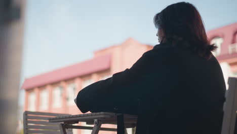 흑색 옷을 입은 여성이 뒷면에 은 빨간 지붕 건물과 함께 야외의 목조 테이블에 놓인 가방을 풀고 있는 근접 측면 시각