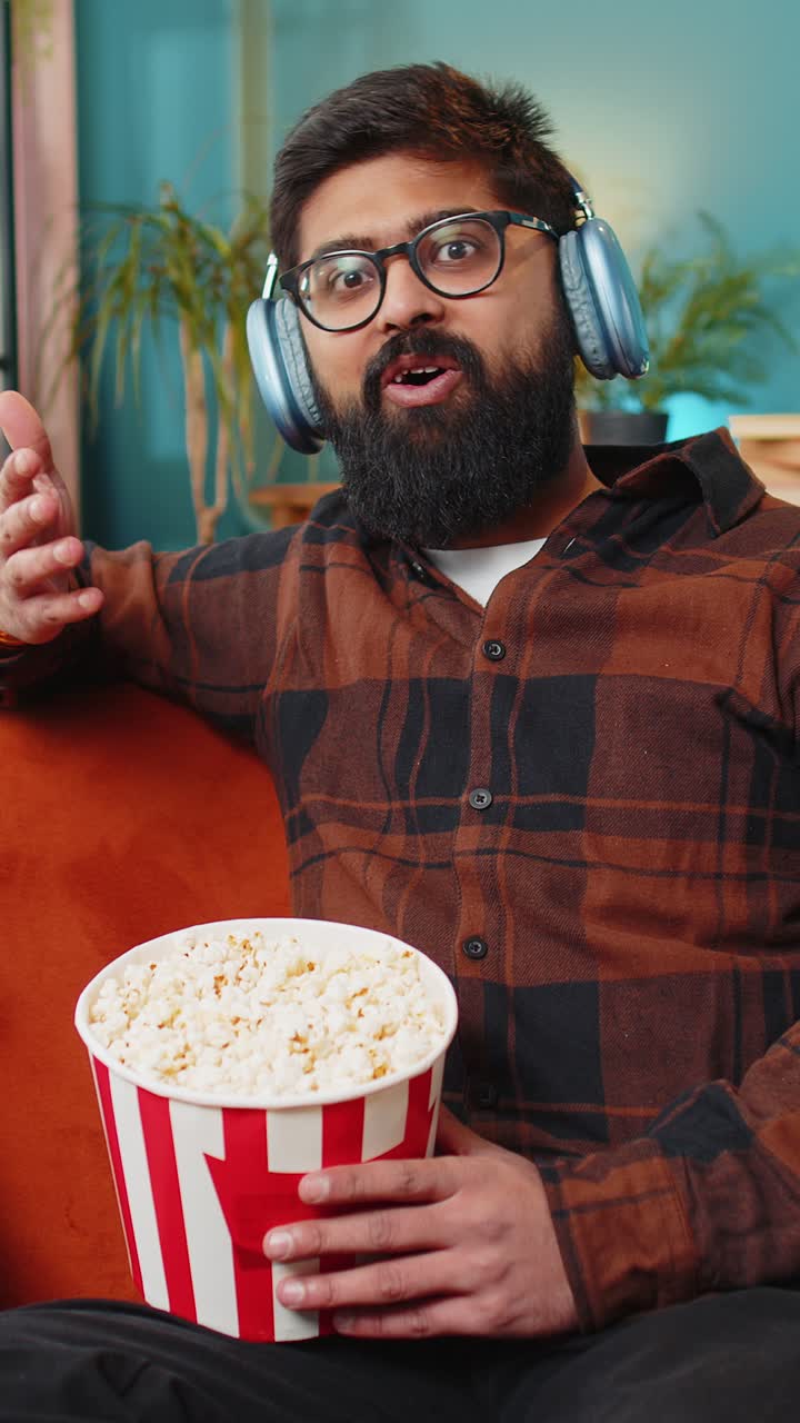 Relaxed indian man eating popcorn watching movie enjoying film during weekend sitting on home sofa