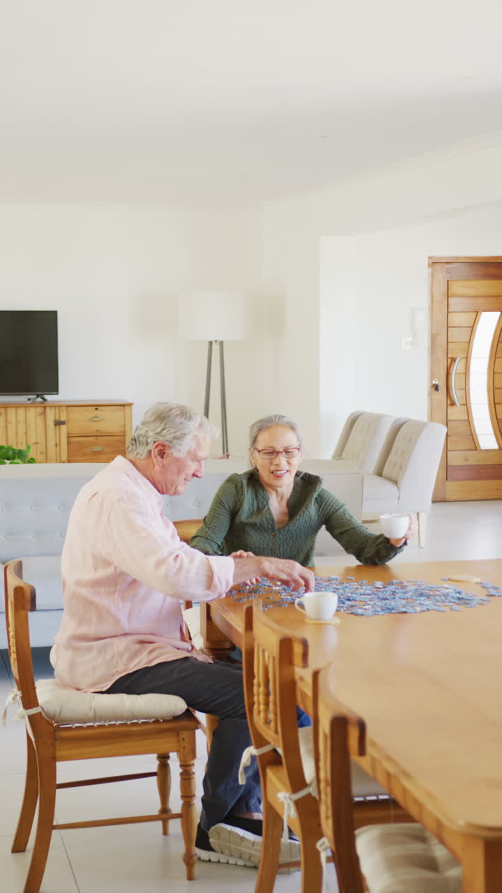 Vertical video of happy diverse senior couple sitting at table and doing puzzle
