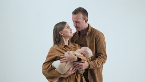 man embracing his beloved wife with sleeping baby in hands. loving couple kissing each other. white backdrop.