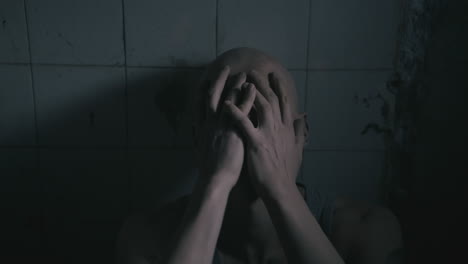 pale bald skinny man sits next to the wall in the creepy old bathroom at night