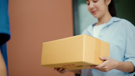 close up parcel in hand of blue uniform delivery man to customer front of the house from shopping online with good service. courier man delivering a cardboard box postal package to destination.