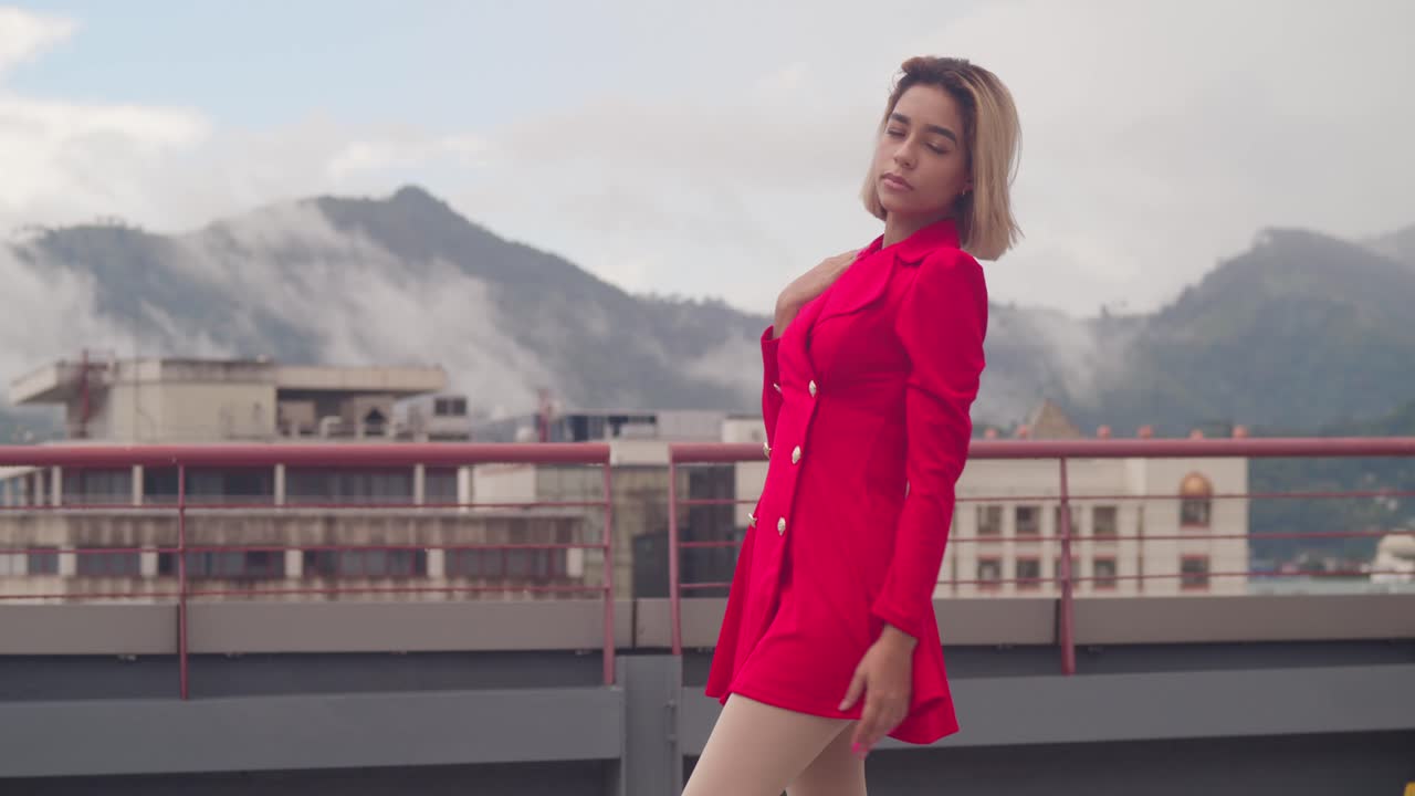 짧은 빨간 드레스가 젊은 히스패닉 소녀를 장식합니다. 그녀는 도시의 지붕에서 바라보고 있습니다.