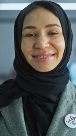 Woman-stands-in-a-modern-polling-station,-poses,-smiles-and-looks-at-camera.-Portrait-of-Muslim-woman,-United-States-of-America-elections-voter.-Background-with-voting-booths.-Concept-of-civic-duty.