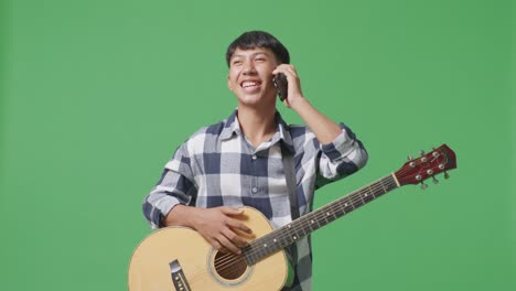 asian teenager with guitar talking on smartphone while standing on green screen background