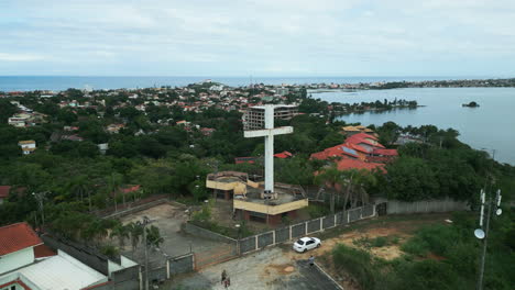 drone moves forward over the white cross at mirante morro da cruz, revealing saquarema lagoon with the city church in the background. saquarema is a venue for the world surf league, wsl.