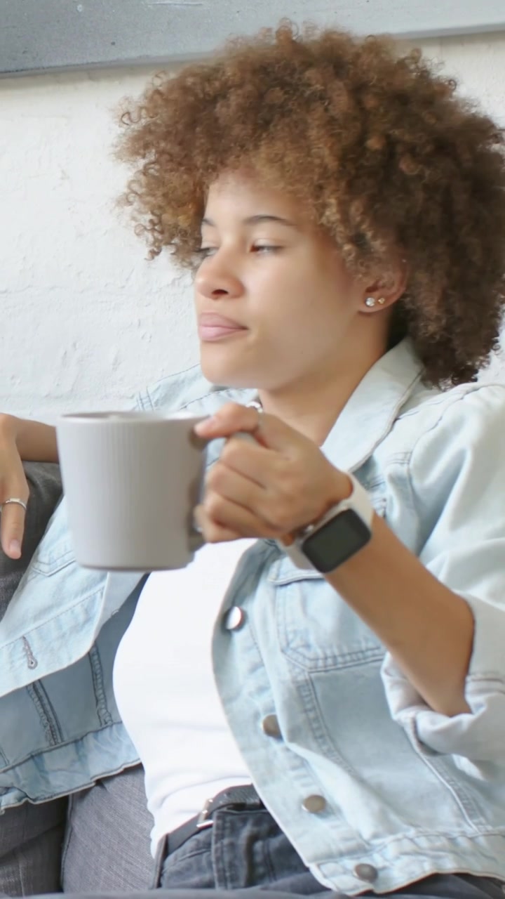 Vertical video: Reaching woman wearing denim jacket noticing ceramic mug on sofa, inhaling scent