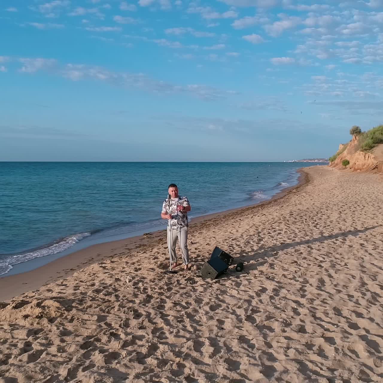 Beautiful sea coast under blue sky. Musician is singing on empty beach with hills near the blue water. Motion camera back