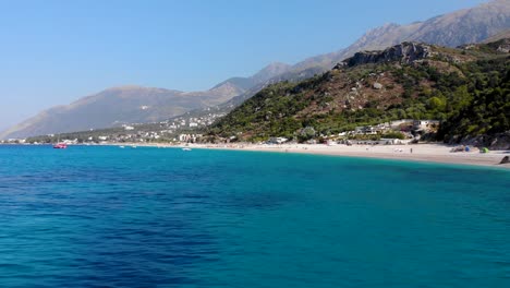 Blue-azure-sea-water-on-rocky-shoreline-of-Mediterranean-with-beautiful-beach-and-mountains-background
