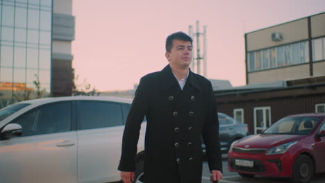검은 코트를 입은 젊은이가 왼손에 브리프케이스를 들고 차 밖으로 나갑니다. 배경에는 주차된 자동차, 사무실 주거 건물, 하늘에 은 구름이 있습니다.