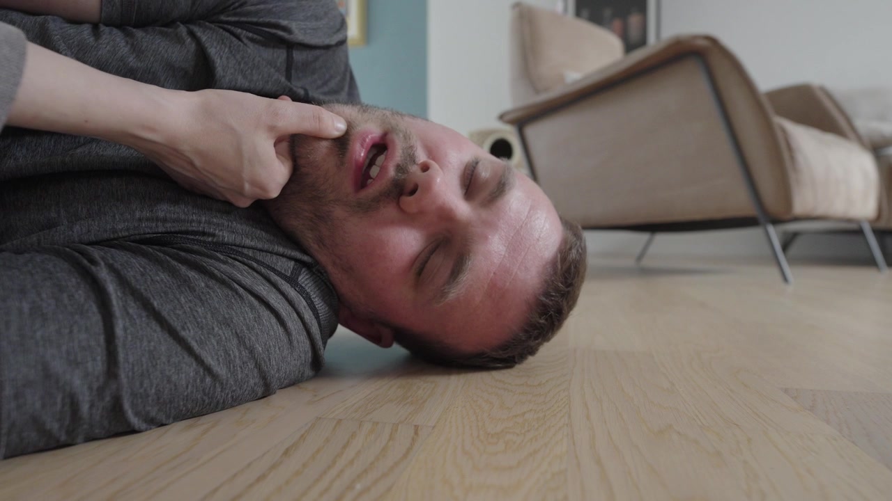 Primo piano di un uomo privo di sensi sdraiato a terra mentre una persona gli controlla il polso e gli apre le vie respiratorie, illustrando la risposta di primo soccorso e una situazione di emergenza a casa.