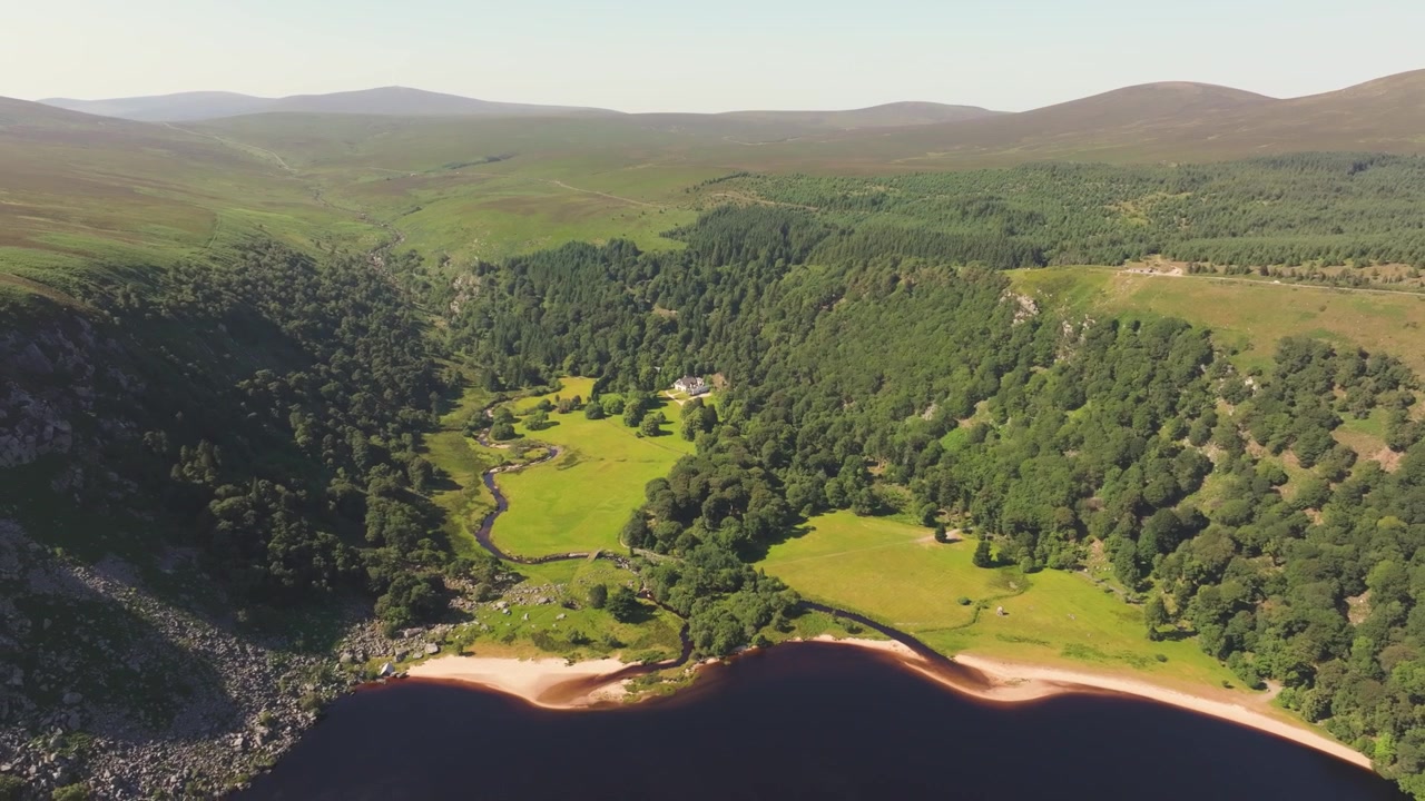 Svingete elv snor seg gjennom lys eng og tett skogkledd dal over en mørkvannsstrand i Wicklowfjellene