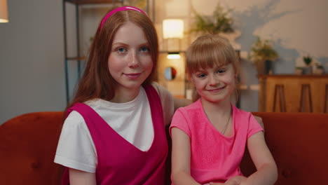 Portrait-of-happy-cute-smiling-two-teenager-girls-friends-siblings-looking-at-camera-sitting-at-home