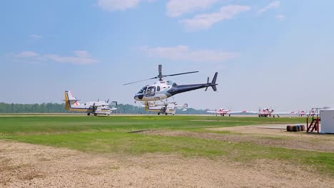 a white helicopter is taking off from a grassy field with small planes and equipment in the background under a clear sky