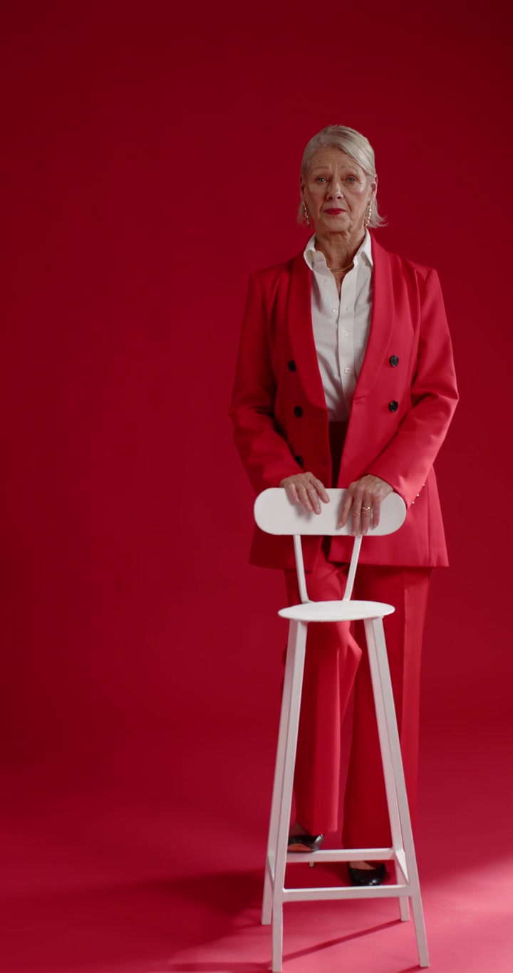 Video verticale: Donna matura in tailleur rosso che tiene uno sgabello in studio, si gira verso la telecamera dopo uno sguardo