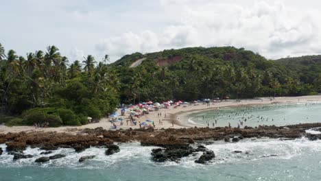 Right-trucking-aerial-drone-view-of-the-popular-tropical-Coquerinhos-beach-covered-in-umbrellas-with-tourists-swimming-in-a-natural-pool-from-a-reef-blocking-small-waves-in-Conde,-Paraiba,-Brazil