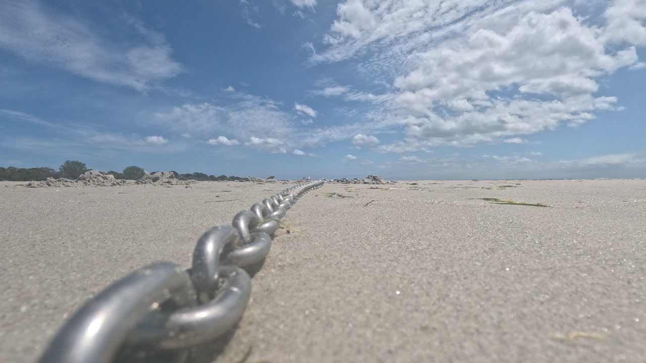 Una catena di metallo giace sulla spiaggia sabbiosa, che conduce verso l'orizzonte mentre le nuvole fluttuano sopra durante il giorno. La scena cattura il legame tra terra e mare