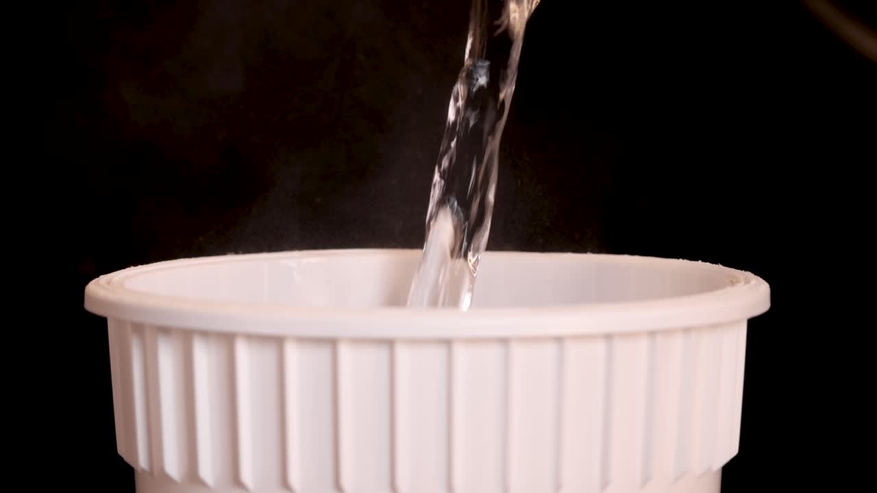 A steady stream of hot water is poured into a white instant noodle cup against a dark background, with visible steam and consistent side lighting