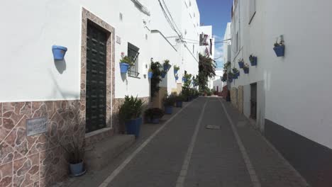 A-view-up-a-narrow-street-with-blue-wall-hanging-flower-pots-in-Conil-de-la-Frontera,-pan-from-left-to-right