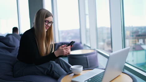 beautiful woman with a phone in office. young businesswoman sitting in a comfortable armchair and rests with a phone near window. laptop and a cup on small table.