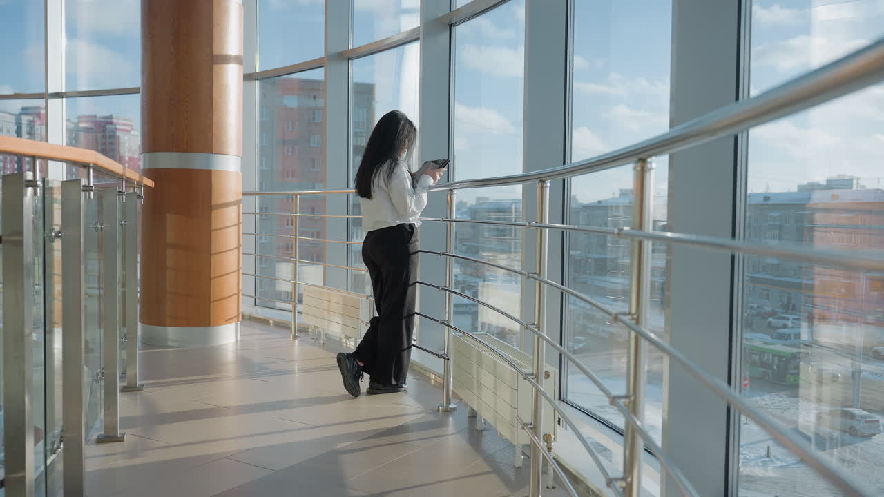 Rear view of professional woman holding tablet walking toward stainless steel railing inside sunlit modern building with large glass windows overlooking urban cityscape on clear winter day