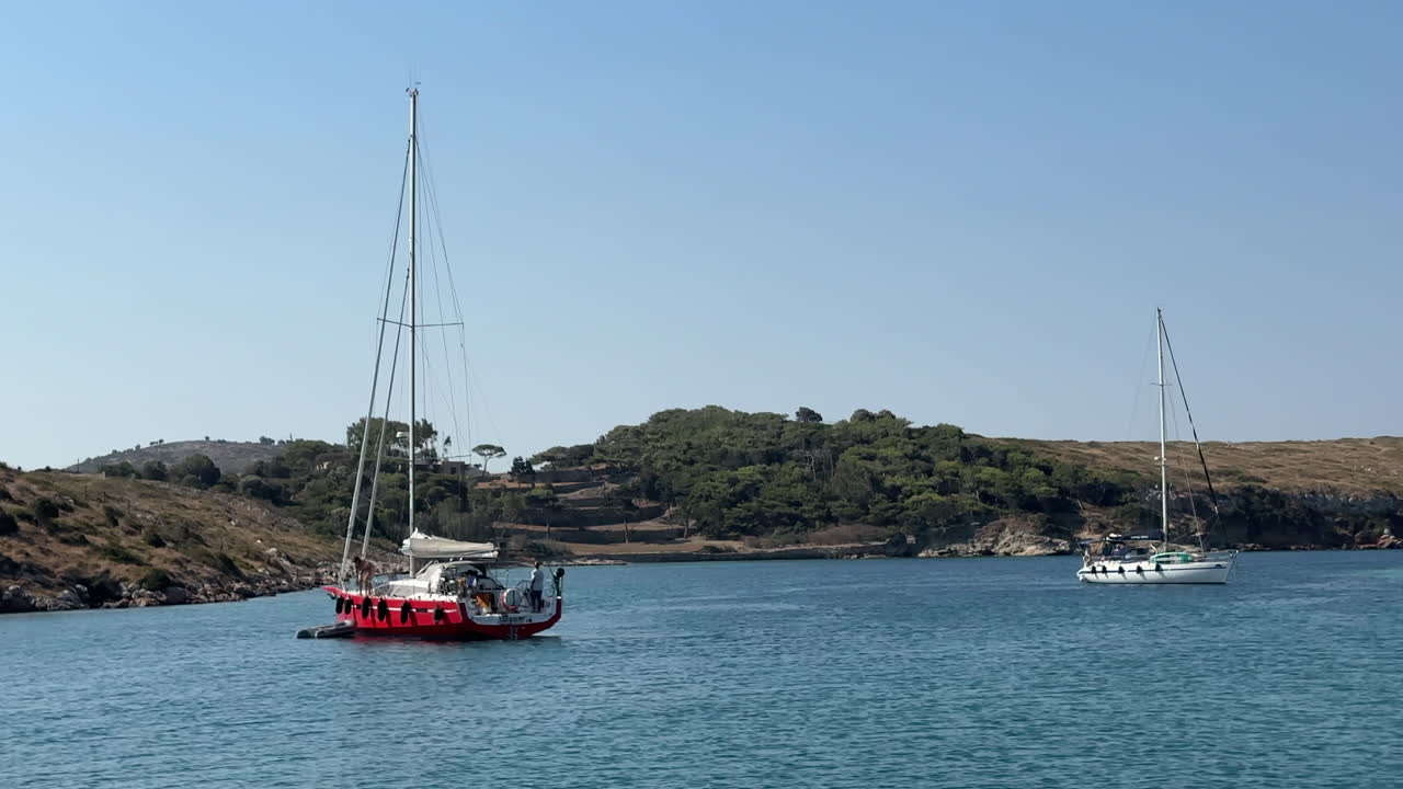 Greece,Arki Island on a calm day, two sailboats on the bay. Red sailboat turning around itself slightly and pulling the dingy out,white sailboat is moving slowly away from the red sailboat.