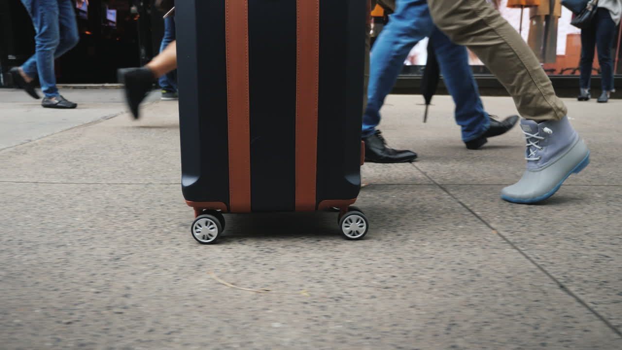 una mujer con una bolsa de viaje sobre ruedas va a un paso de peatones en una multitud de personas que solo tienen piernas