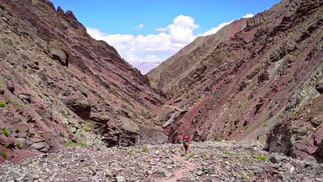 Man-hiking-in-a-dry-deserted-valley,-a-backpacker-walking-away-from-the-camera-on-a-stony-road-between-high-mountains-during-the-day