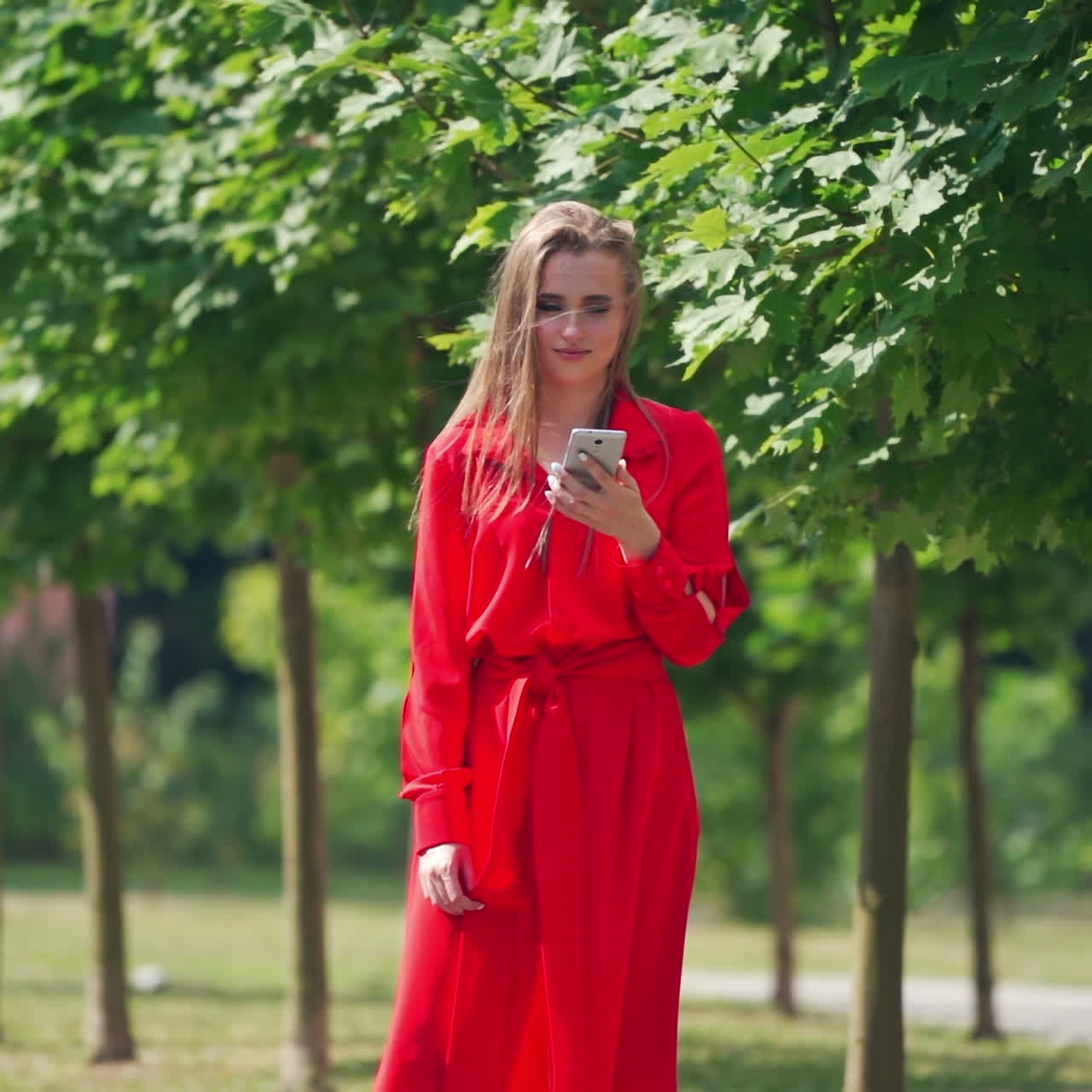 Lovely model with a cellphone. Beautiful young woman in bright red dress with a phone on the green trees background in summer. Slow motion.
