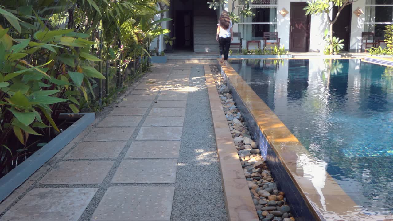 Young Lady Walking Along the Edge of a Swimming Pool in a Tropical Hotel Shot From Behind