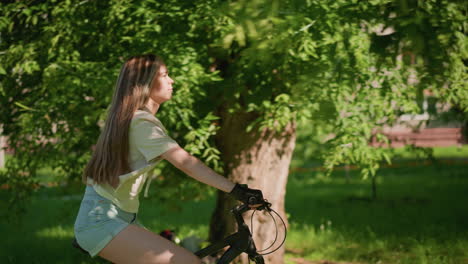 vista lateral de una mujer joven que anda con confianza en bicicleta a través de un parque verde exuberante, su largo cabello fluye detrás de ella, expresando una determinación tranquila, el fondo muestra vegetación y sombras suaves