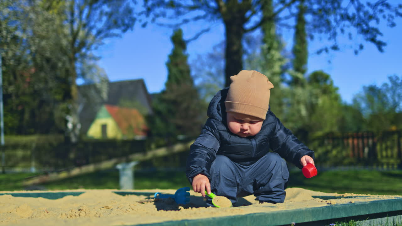 Little two-year-old boy plays with sand outside in a sandbox. Toddler boy digs sand with a shovel on a playground. Child's walk outdoors