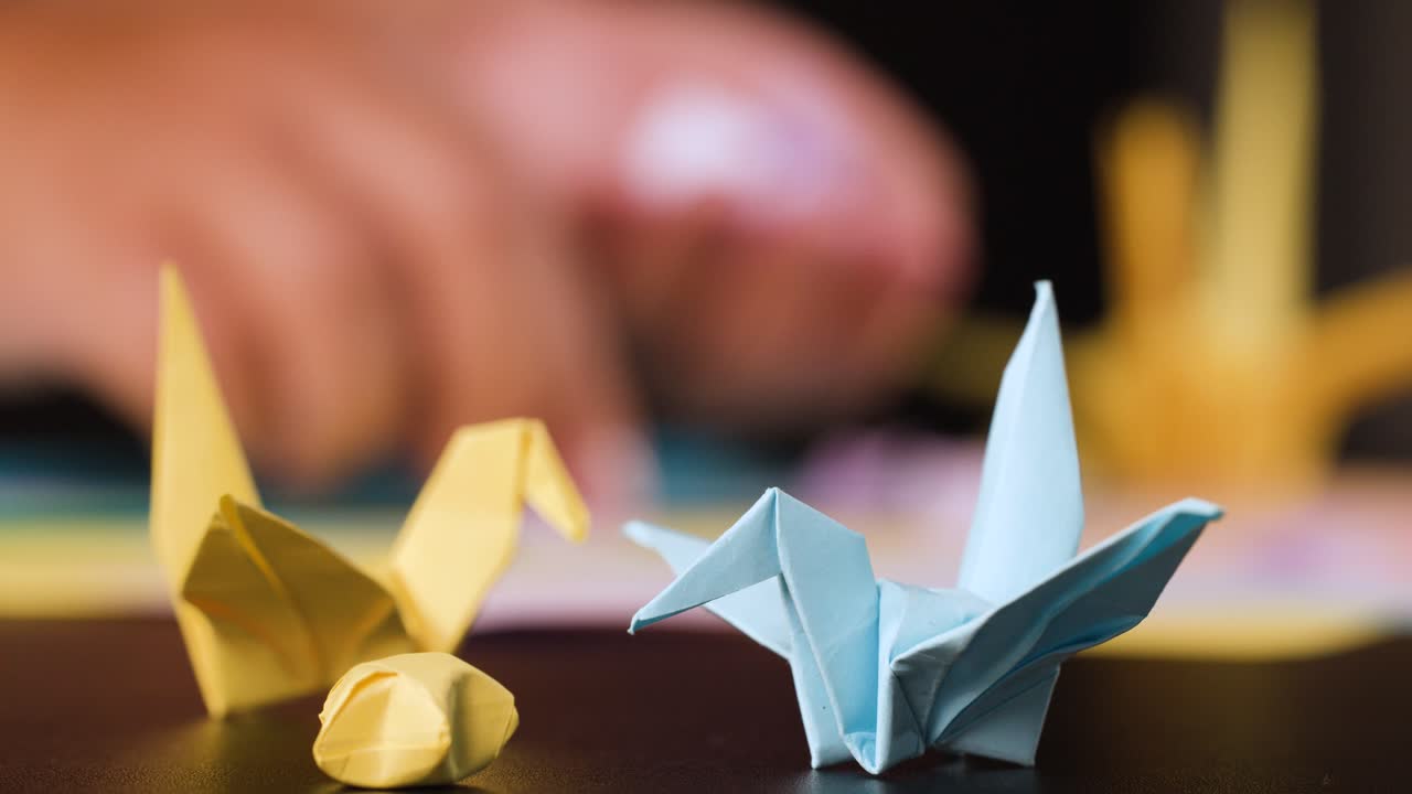 Primo piano di mani che piegano la carta dietro a degli origami a forma di gru completati, di colore giallo e blu