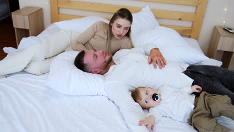 caucasian parents and their little baby lie on the big double bed. little toddler with pacifier in mouth tries to get up on his feet.