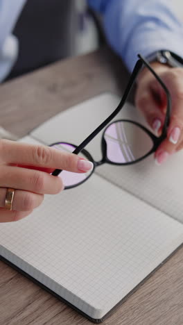 Female hands holding the glasses. Woman puts off the glasses and closes her paper notebook. Close up. Vertical video