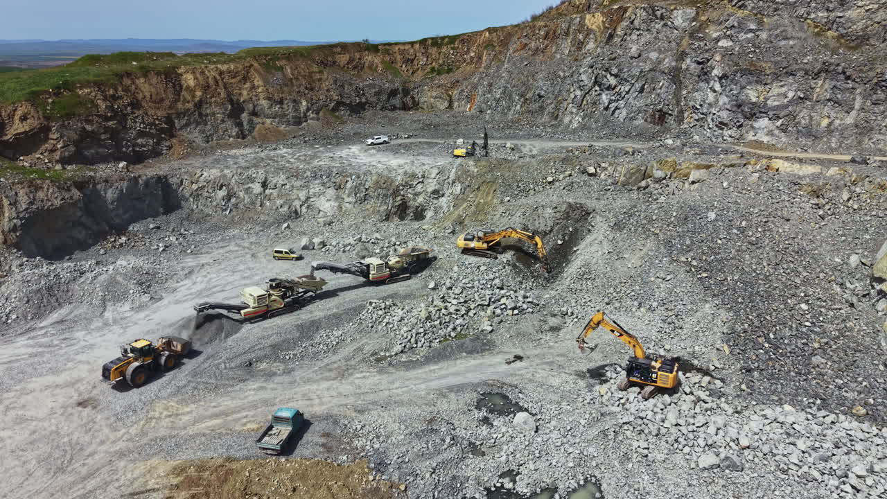 Work takes place at a granite quarry with heavy equipment moving stone and rock in a large open area under a clear sky