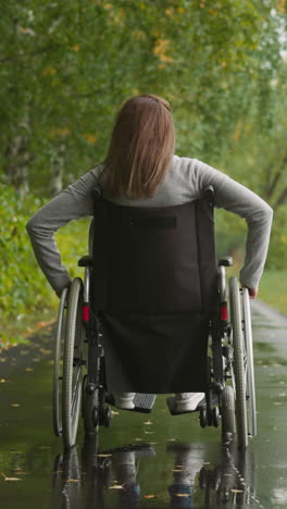 mujer joven en silla de ruedas camina en el parque bajo una lluvia torrencial. mujer gira las ruedas moviéndose a lo largo de un camino mojado. rehabilitación física después de una lesión vista trasera