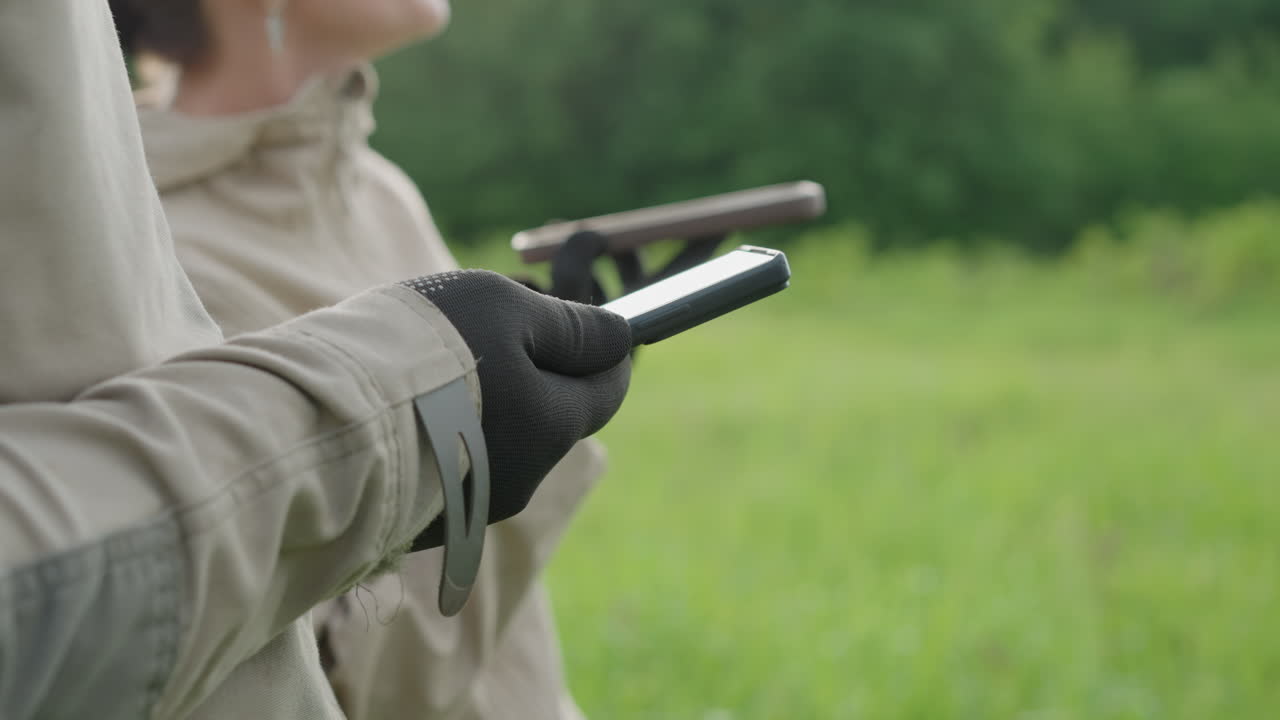 close up adult wearing dotted glove holding touchscreen phone in vast green meadow, partial view of companion gesturing and monitoring device, blurred tree line backdrop