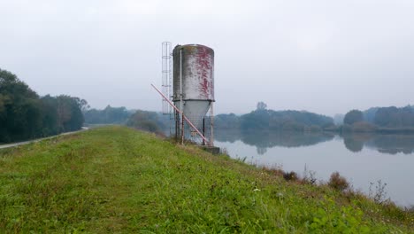 Distant-drone-shot-of-an-old-metal-silo-overlooking-a-reflective-lake-on-an-autumn-morning
