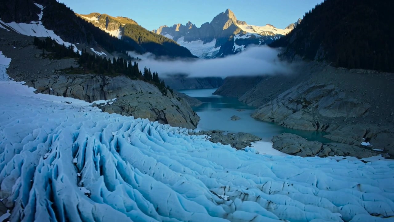 Drone con telecamera che si lancia, scivolando per catturare crepacci glaciali, nebbia su un lago turchese e cime illuminate dal sole