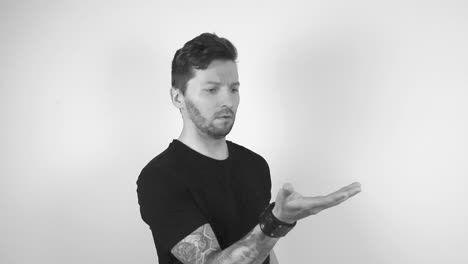 grayscale footage of a tattooed man with short hair and trimmed beard lifting up his hand with nothing on it, then he is surprised and looks into the camera with a stunned look on his face.