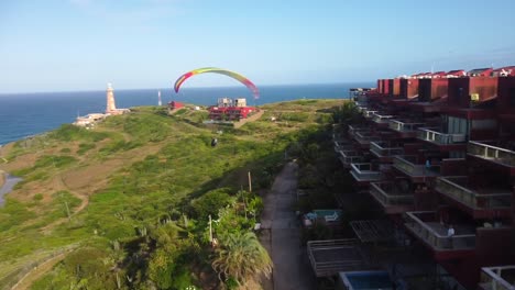 Paragliding-over-a-coastal-town-with-a-lighthouse-and-lush-green-hills