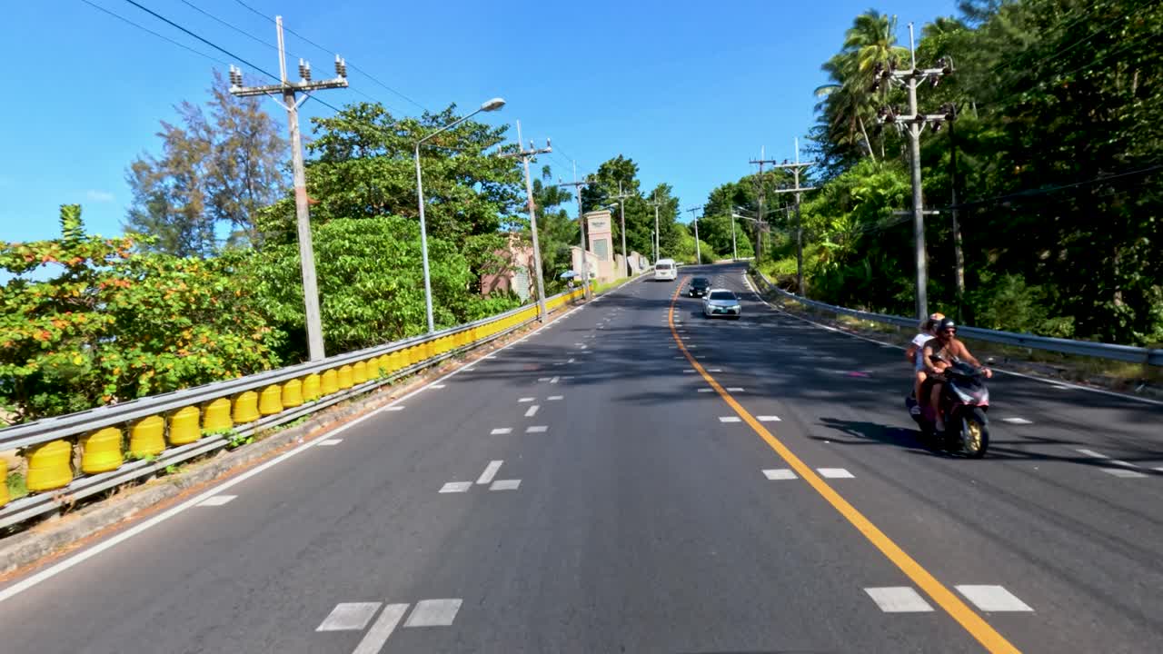 Vehicle travels Phuket road, passing palm trees, cars, and motorbikes under bright daylight