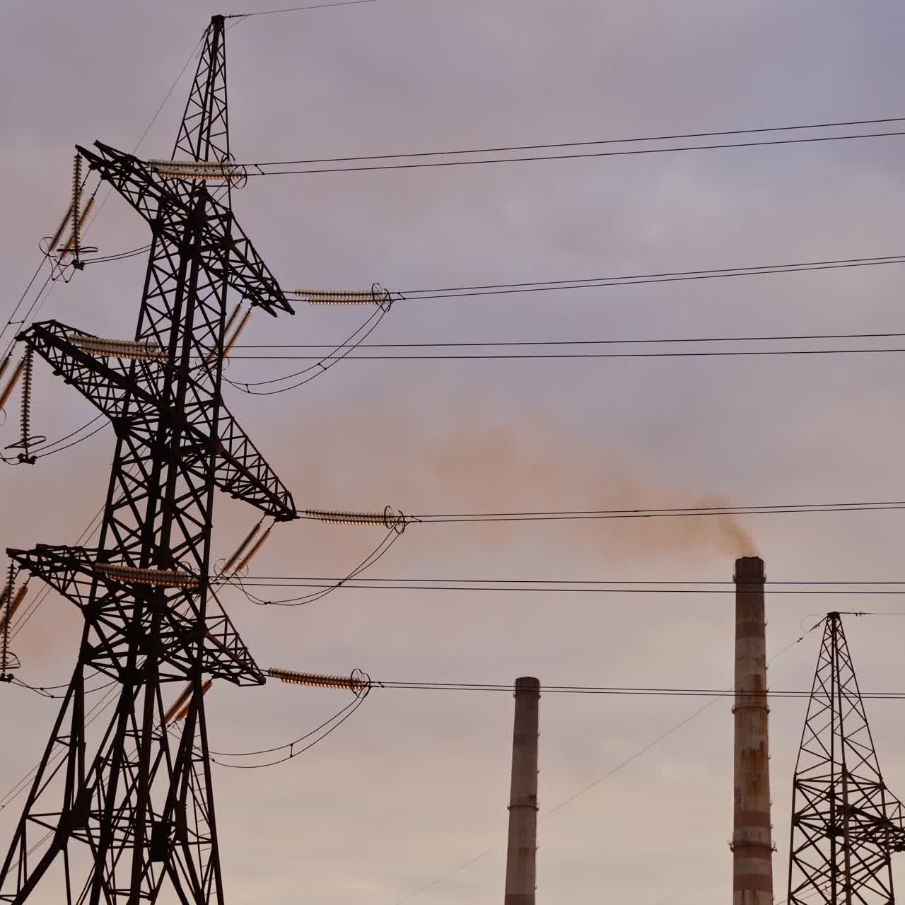 Electric towers on factory pipes background. High-voltage electric line for current distribution. Steel pylons with wires in the evening.
