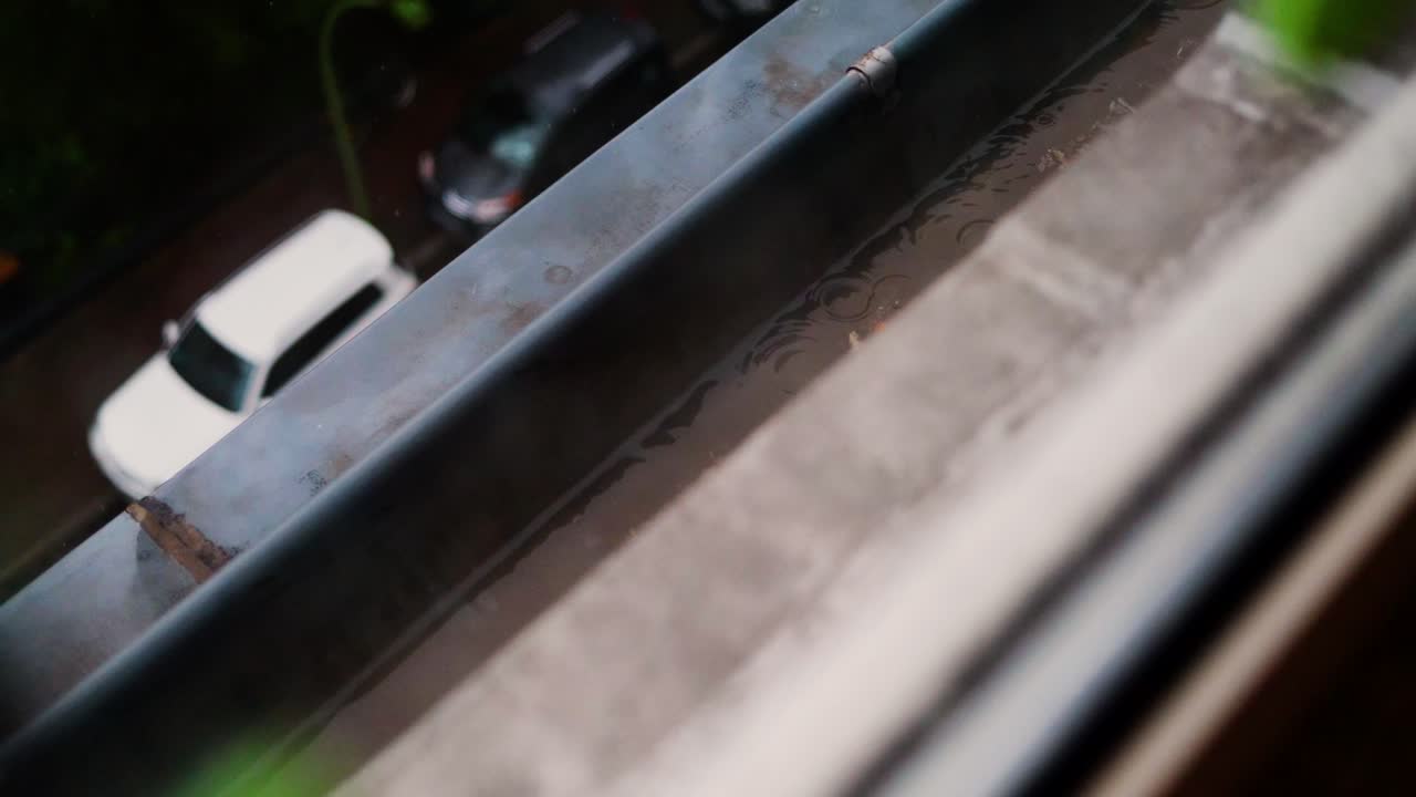 Rain falling on a windowsill with blurred view of parked cars below on a gloomy day