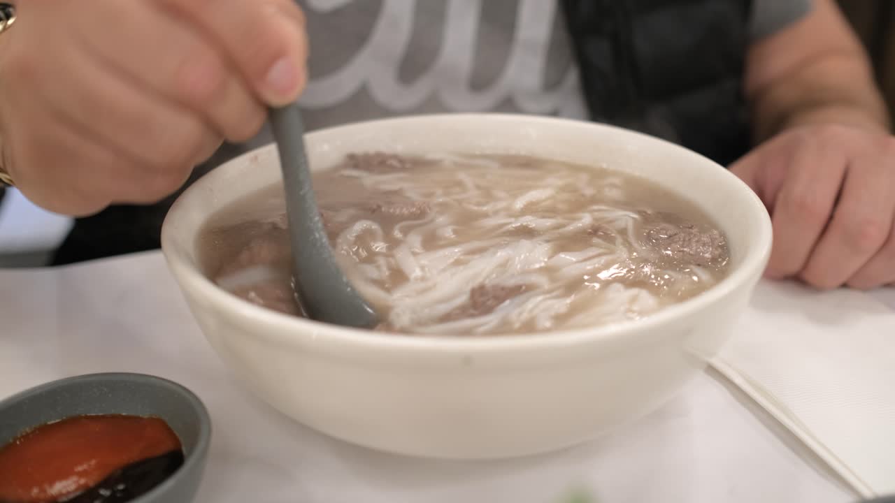 Pria muda mengaduk sup pho-nya dan menambahkan saus siracha serta hoisin.