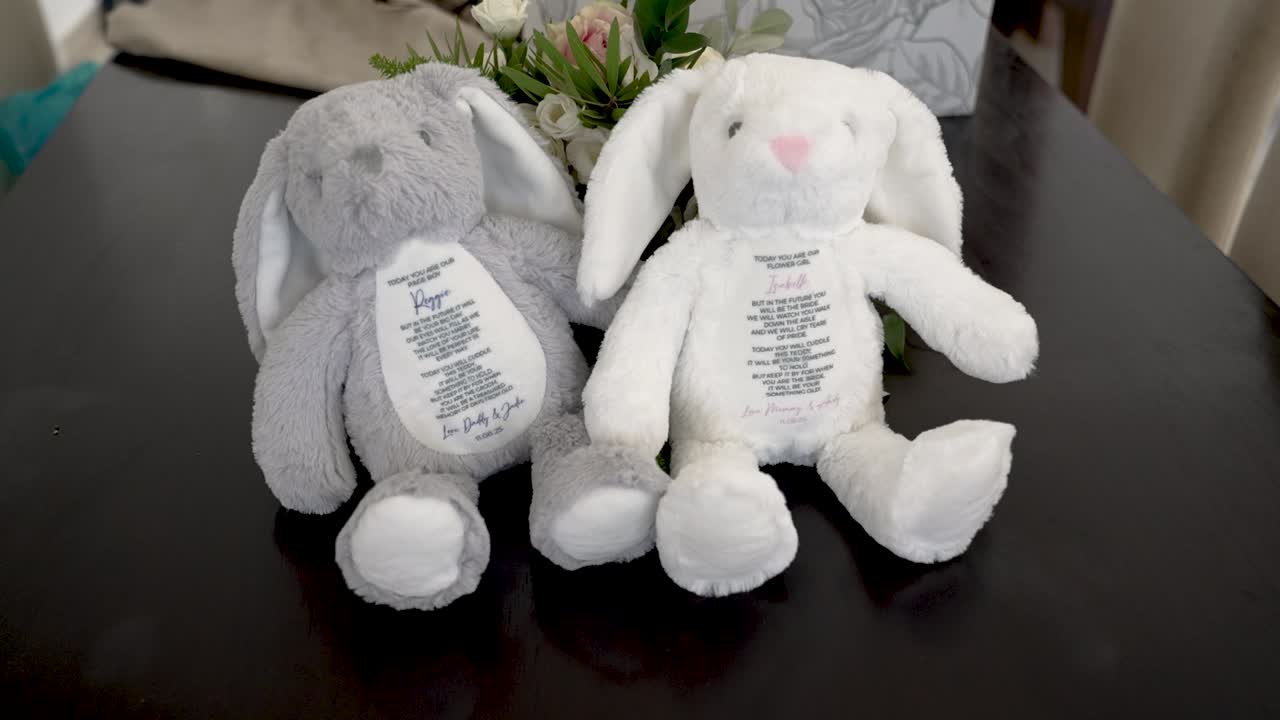 Two plush bunnies sit on a table, one gray and one white, each featuring a special message sewn on their tummies. They make perfect gifts for loved ones