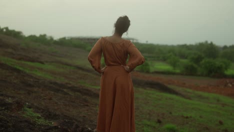 A-woman-in-a-brown-dress-stands-on-a-grassy-hillside-looking-into-the-distance